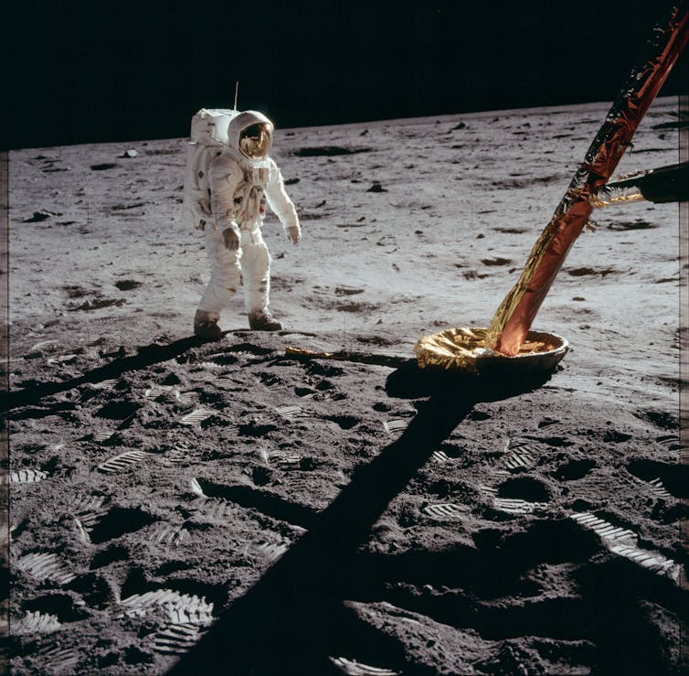 An astronaut looks at part of a landing apparatus, while some golden light is reflected on their helmet.